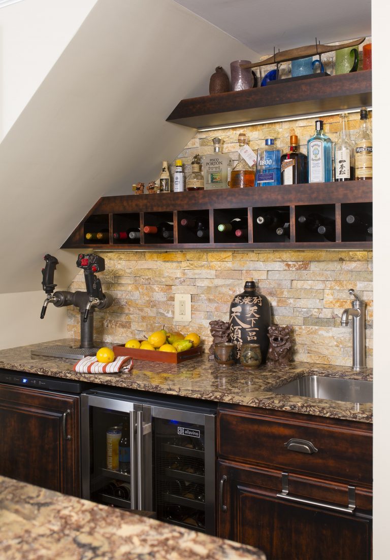 home basement bar under stairs warm color tones dark cabinets stone backsplash open shelving and storage beverage refrigerator